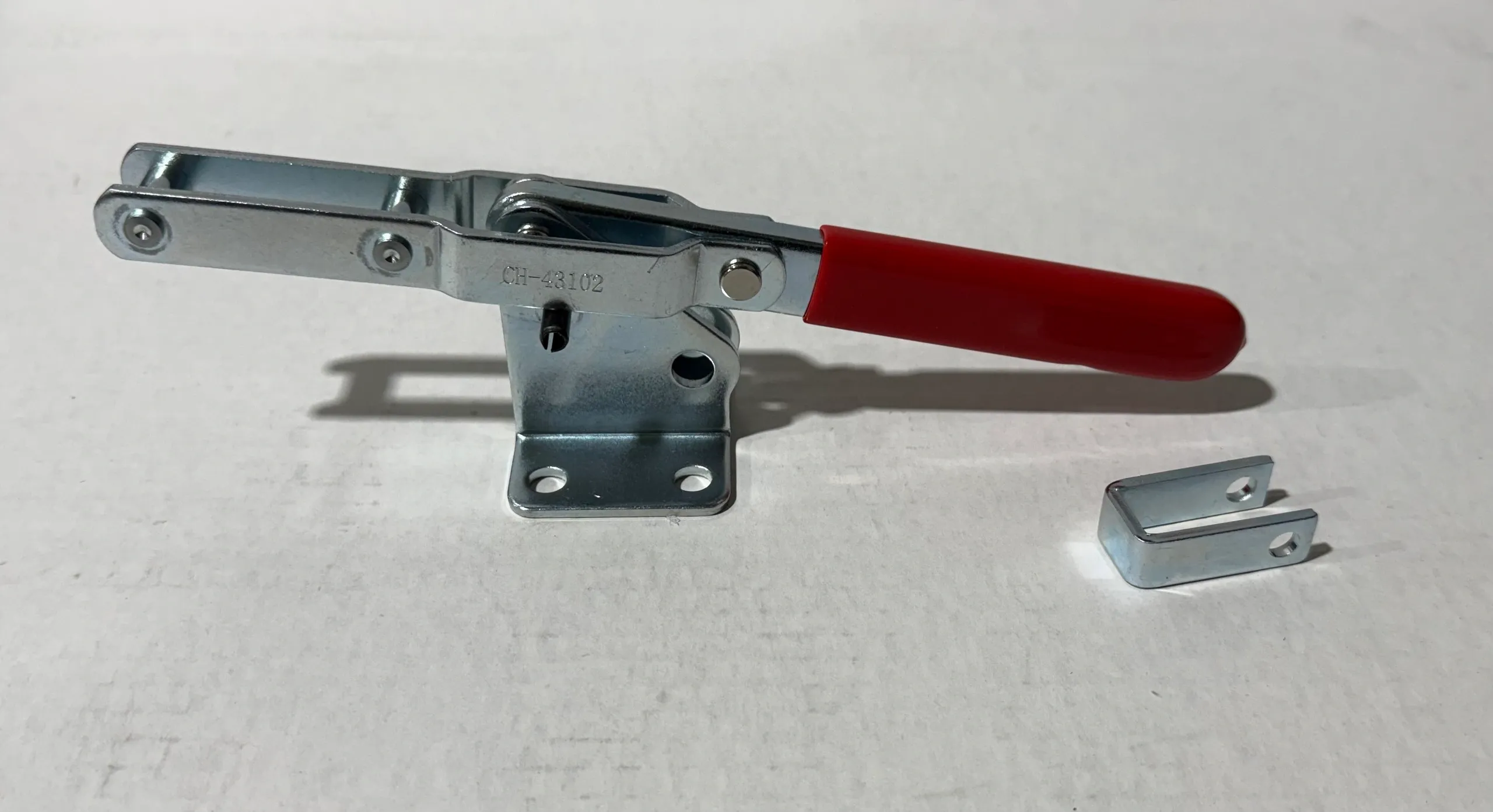 A metal toggle clamp with a red handle on a white surface.