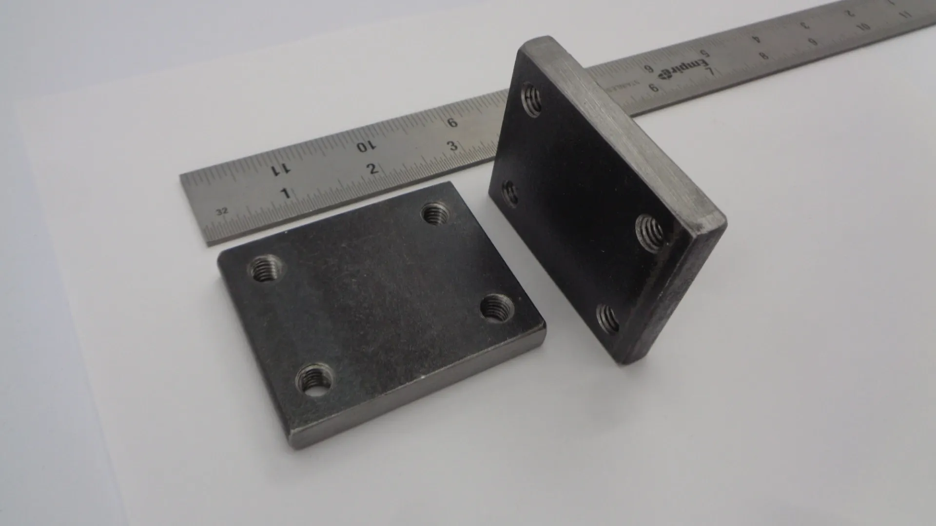Two black metal flange plates with holes next to a ruler.